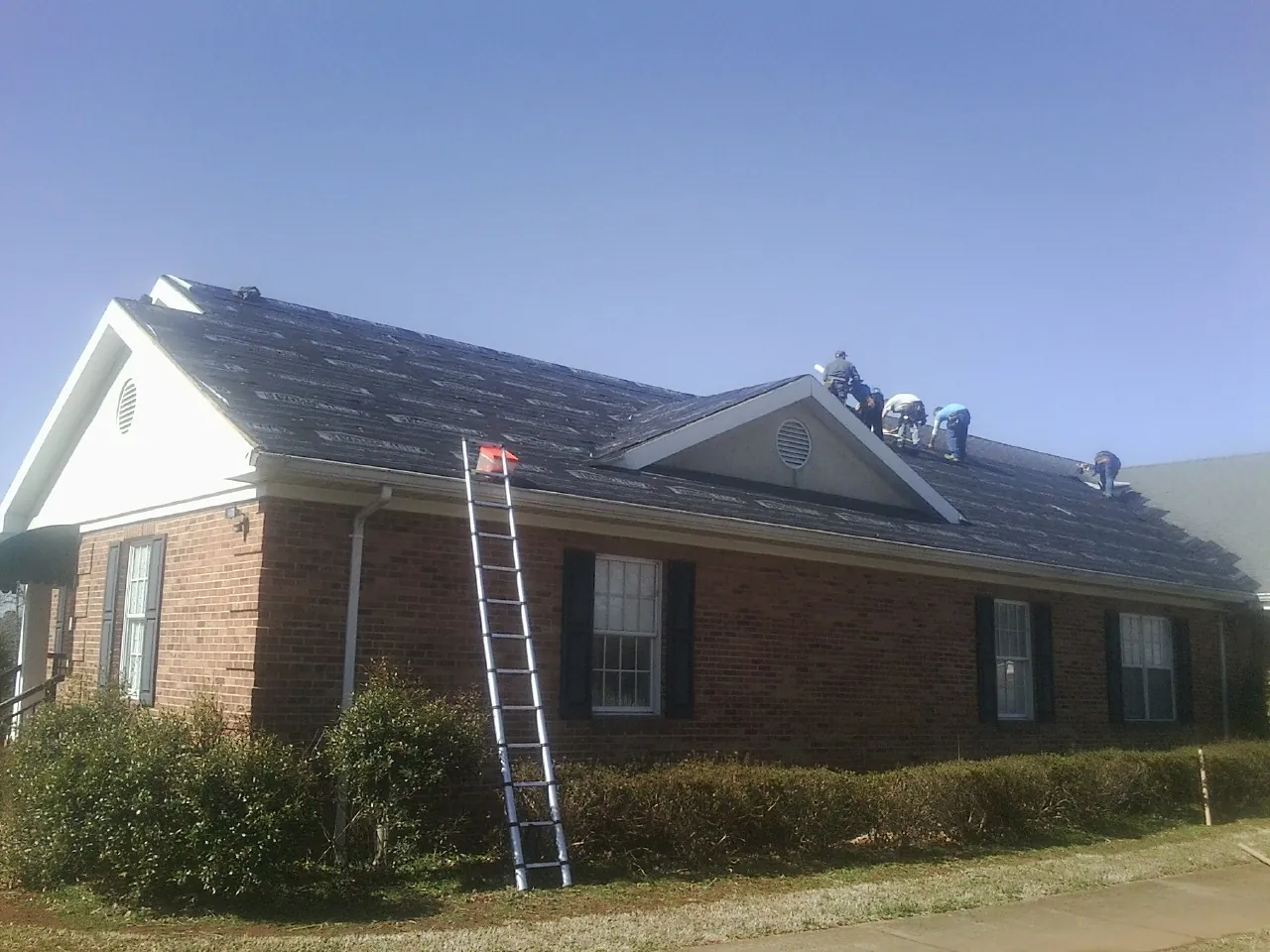 Crew tearing shingles off a brick ranch roof with a ladder set — Metal Roof Repair work by our crew in Bosque Farms