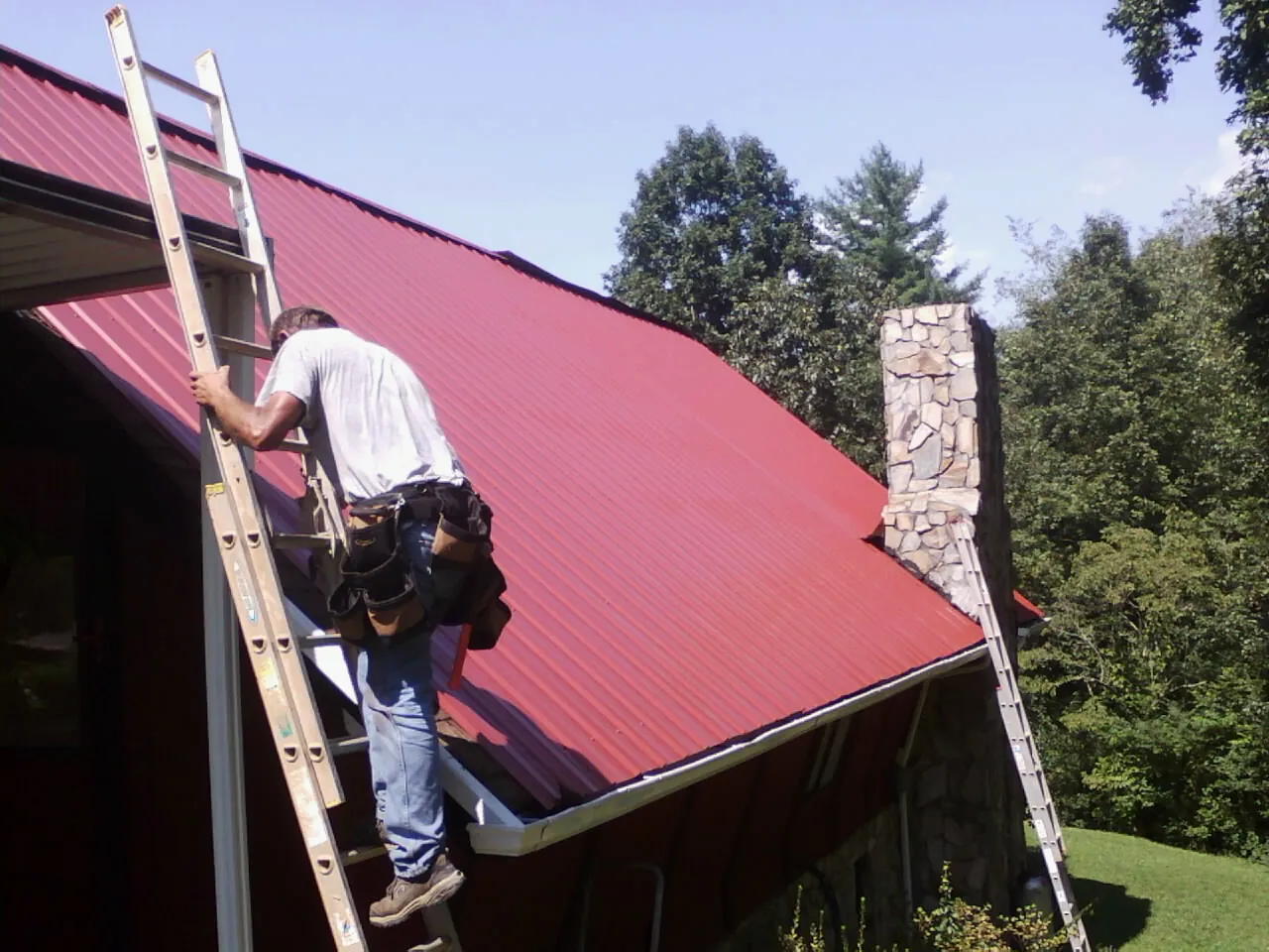 Roofer on a ladder beside a red metal roof and stone chimney — Soffit & Fascia Repair by Roofing Brothers in Bosque Farms